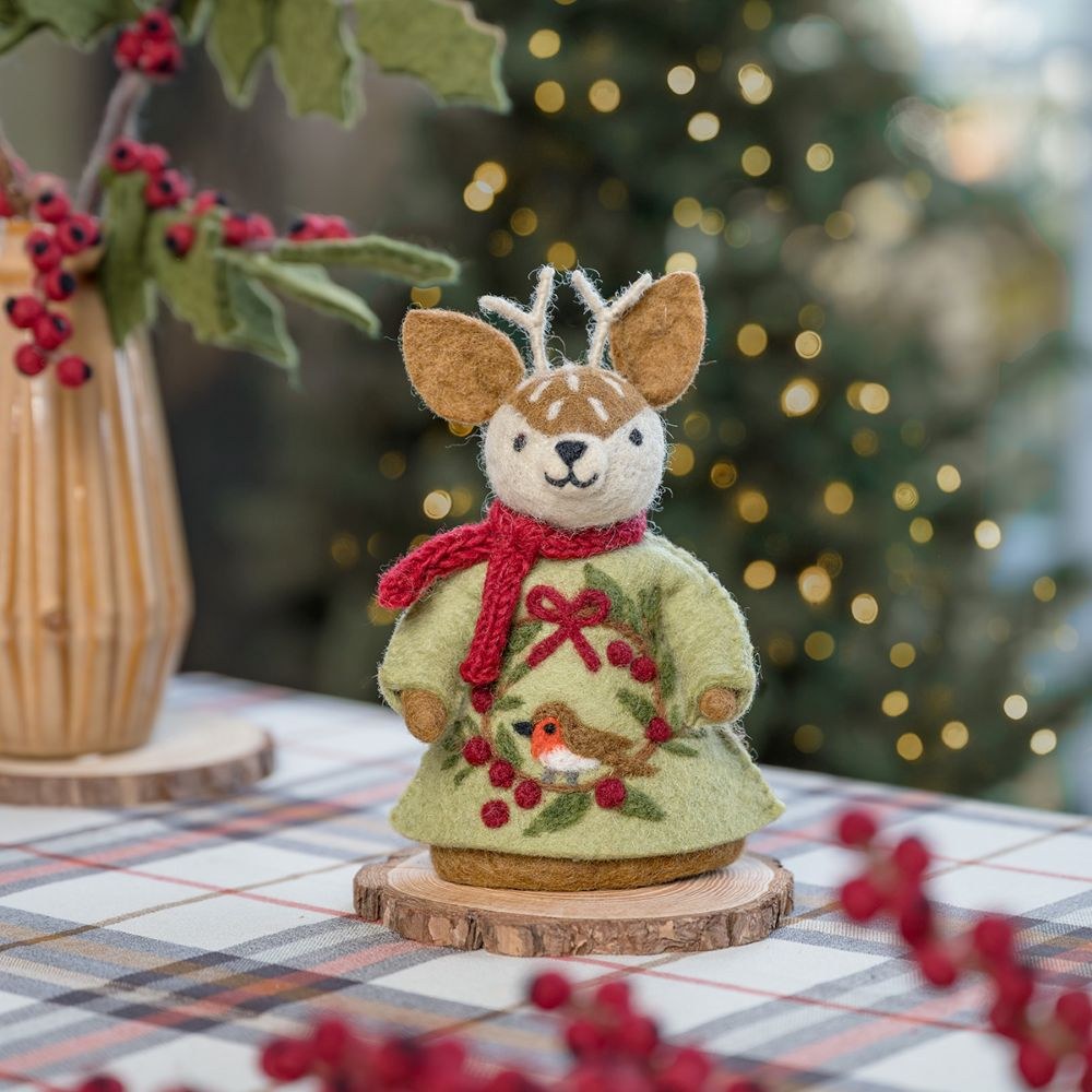 Sjaal met Verhaal vilten hertje in groen jurkje met rood sjaaltje, staand op houten schijf tussen rode bessen en kerstdecoratie